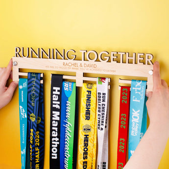 Wooden medal holder with 'Running Together' text, personalised with two lines of custom text being held up to a wall before installing