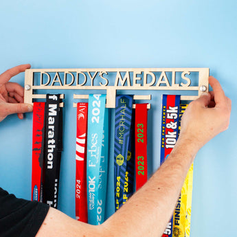 Personalised Minimal Wooden Medal Hanger featuring 'DADDY'S MEDALS' being positioned before installation, using the included Brushed Aluminium Wall Mount Fixings