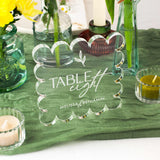 Clear glass table number with 'Table eight' and 'Melissa & Benjamin' text, placed on a green tablecloth with candles and flowers.