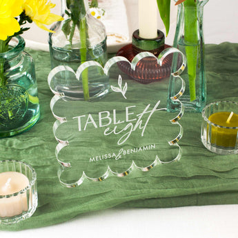 Clear glass table number with 'Table eight' and 'Melissa & Benjamin' text, placed on a green tablecloth with candles and flowers.