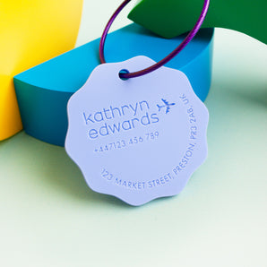 A purple luggage tag with a wavy circle design and an engraved aeroplane. It has personalised text including a name and contact details.